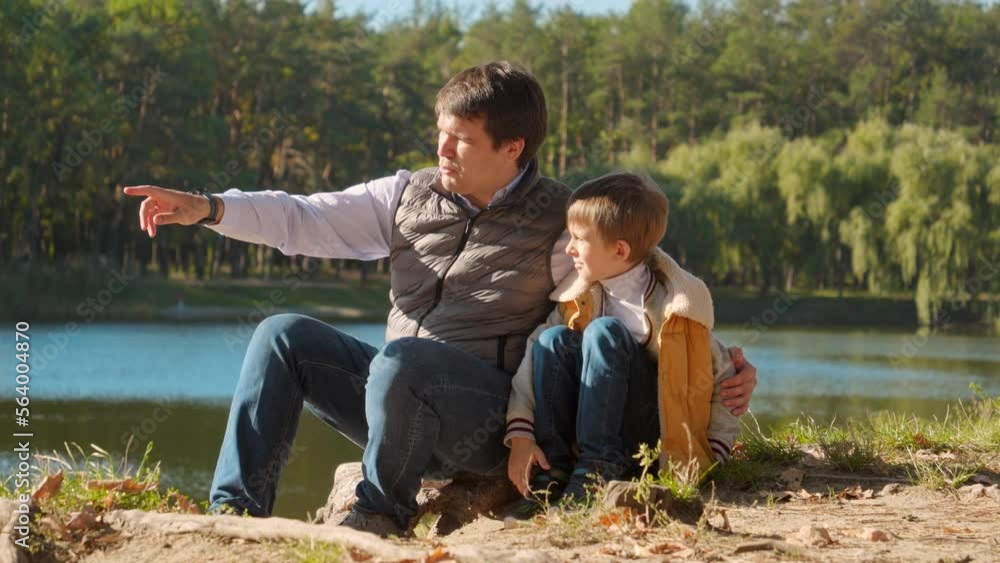 Young caring father talking to his son while sitting at the lake in forest. Family outdoors, happy parenting and childhood, autumn landscape, kids and parents relaxing outside Young caring father talking to his son while sitting at the lake in forest. Family outdoors, happy parenting and childhood, autumn landscape, kids and parents relaxing outside