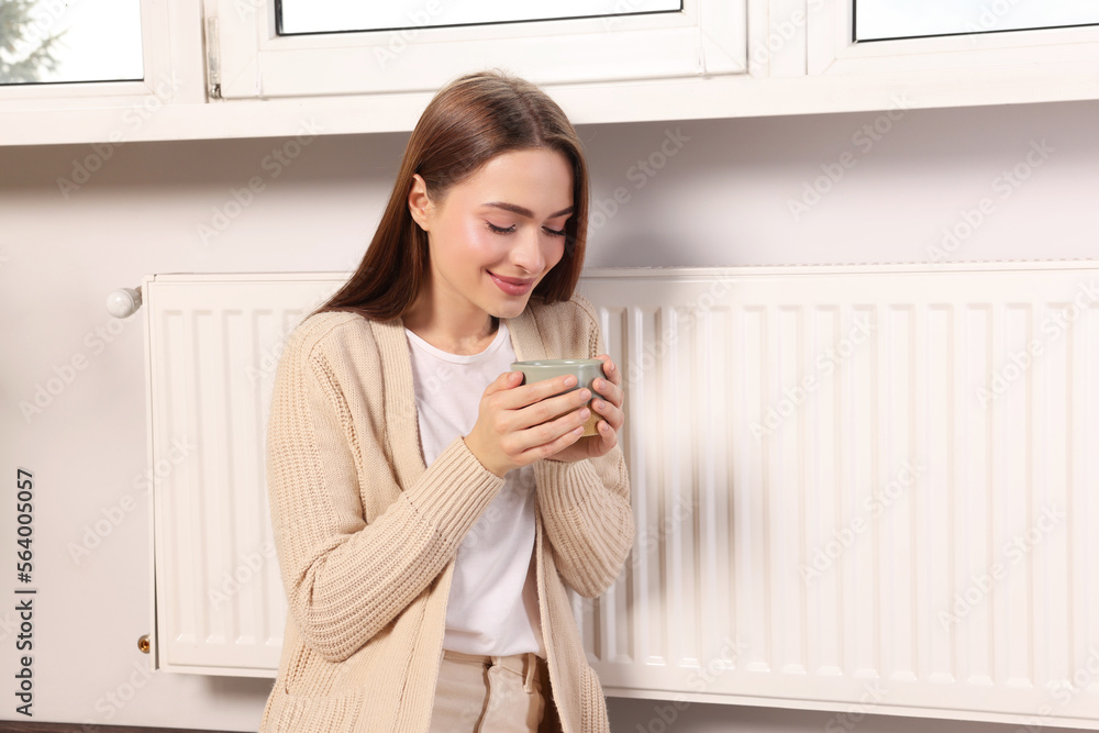 Woman holding cup with hot drink near heating radiator indoors