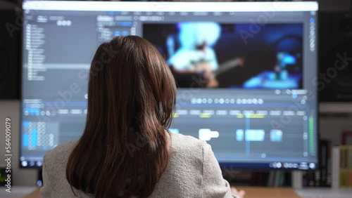 Back view of female video editor editing new project film montage on full screen computer sitting at office or home. Content creator in digital company processing movie with post production software
