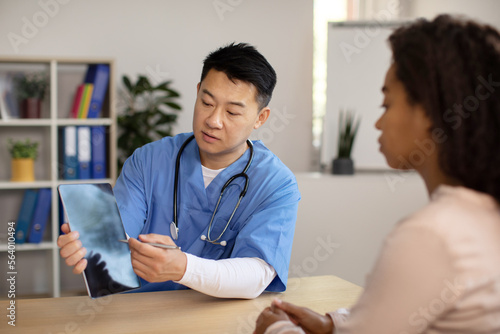 Serious middle aged korean doctor in uniform consults young african american lady, shows tablet with blank screen