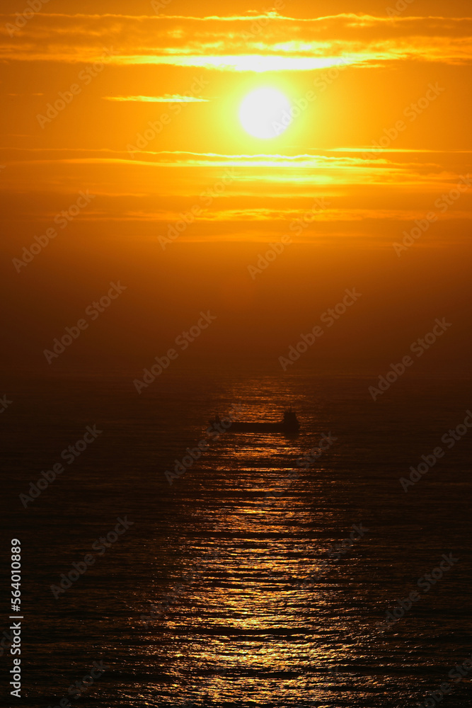Bulk cargo ship passes through Cabo de Peñas during sunset in contra light with the sun in the background