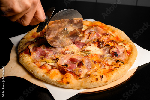 hand gently cuts with a pizza cutter round pizza with bacon on wooden chopping board
