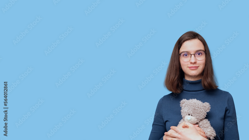 Portrait of a beautiful student girl with glasses, hugging a gray teddy bear toy on a blue background