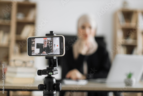 Focus on screen of phone recording video blog of beautiful muslim woman in hijab talking while looking at modern smartphone. Female blogger sitting at home and doing live stream.