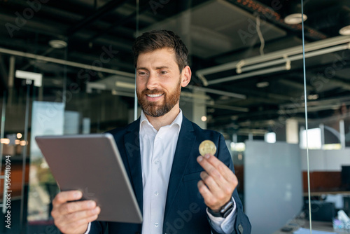Businessman looking at tablet and holding bitcoin coin, analyzing financial data at workplace in office interior