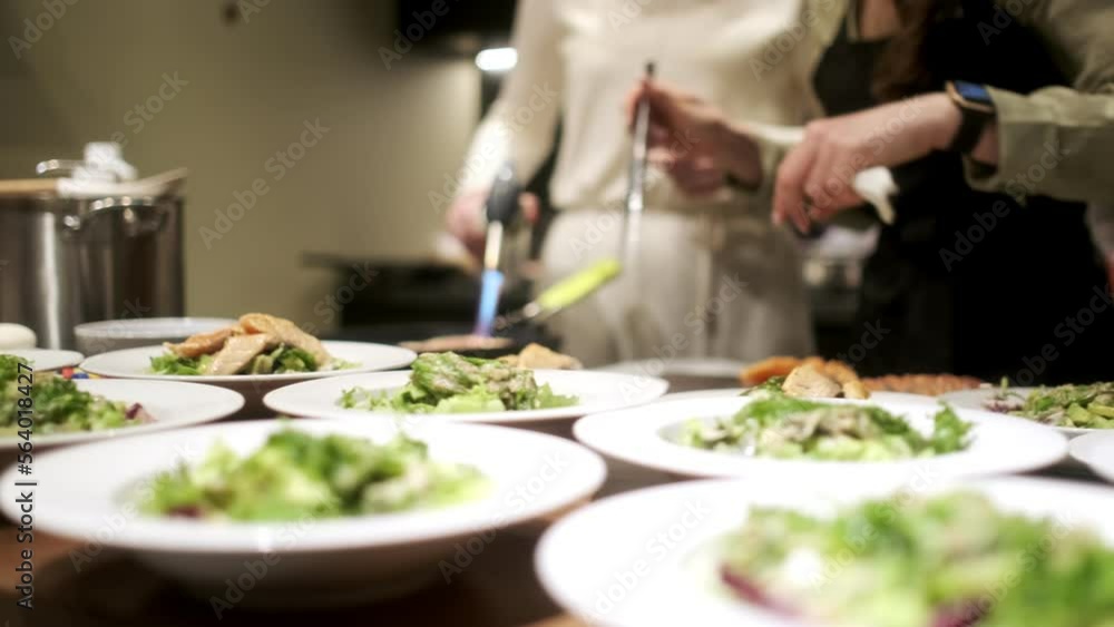 One chef roasts salmon with a gas burner. The second arranges the finished salmon on plates. In the foreground are plates of salad for a group of people. The concept of cooking dinner. One chef roasts salmon with a gas burner. The second arranges the finished salmon on plates. In the foreground are plates of salad for a group of people. The concept of cooking dinner.
