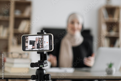 Charming muslim woman in hijab and headset talking and gesturing while recording video on modern phone. Female blogger sitting at home and doing live stream.