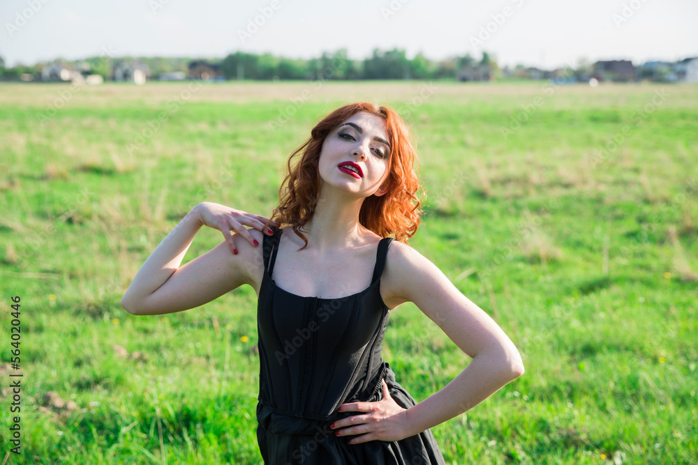 A bright red -haired girl with a bright red lipstick in a black dress in the midst of a field in a sunny control light. Selective focus. A bright red -haired girl with a bright red lipstick in a black dress in the midst of a field in a sunny control light. Selective focus.