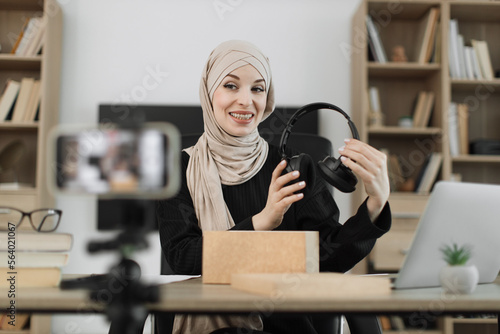 Arab woman recording video on phone camera while unpacking box with new wireless headphones. Female influencer sharing with subscribers her positive feedback about new order.
