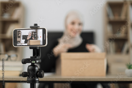 Excited smiling muslim woman in casual wear and hijab recording video on camera while unpacking gift boxes. Female blogger sharing her emotions with her subscribers in social networks.