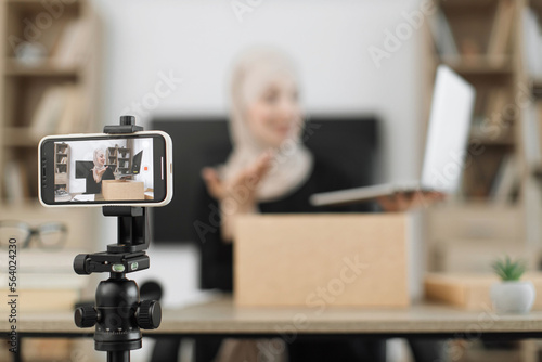 Focus on phone screen , muslim woman recording video on phone camera while unpacking box with new wireless laptop. Female influencer sharing with subscribers her positive feedback about new order.