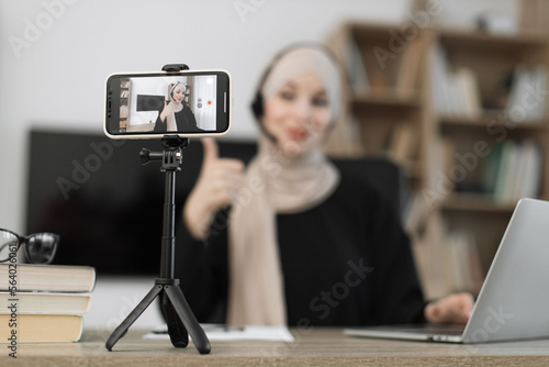 Focus on screen of smartphone of attractive young muslim woman in hijab and headset taking selfie on her smart phone using stick. Female blogger doing live stream showing thumb up.