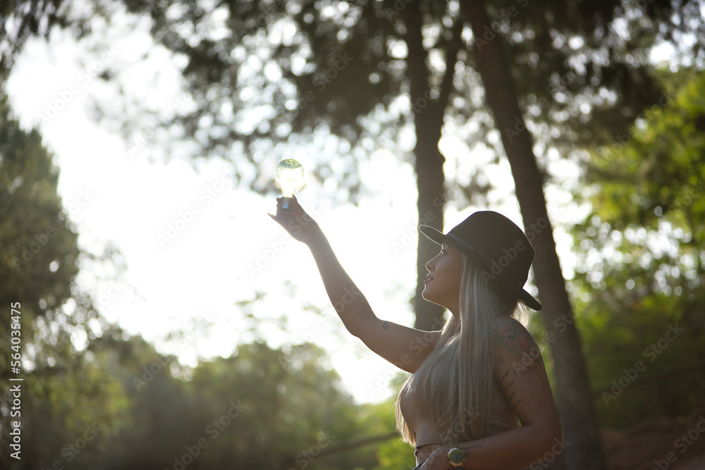 Naklejka premium Young, beautiful, blonde woman with hat, holding a light bulb illuminated by the sun passing through it. Concept light, energy, electricity, renewable, solar.