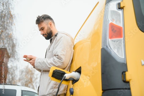 bearded caucasian man standing near an electric car that is charging and making time adjustments on a smartphone.