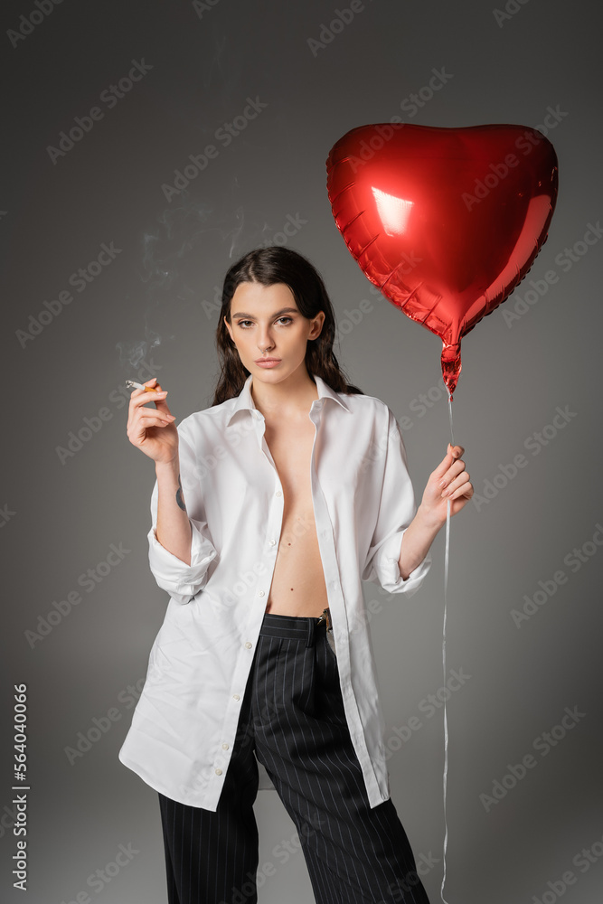 trendy woman in white unbuttoned shirt holding cigarette and red heart-shaped balloon while looking at camera on grey. trendy woman in white unbuttoned shirt holding cigarette and red heart-shaped balloon while looking at camera on grey.