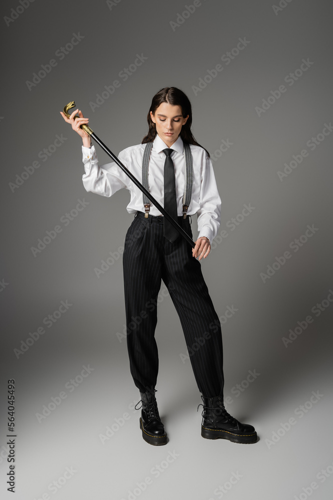 full length of stylish woman in elegant oversize outfit and laced-up boots holding walking cane on grey. full length of stylish woman in elegant oversize outfit and laced-up boots holding walking cane on grey.