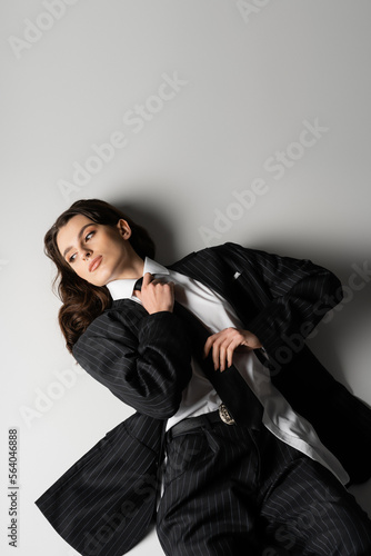 high angle view of brunette woman in black oversize suit touching tie and looking away while lying on grey background.