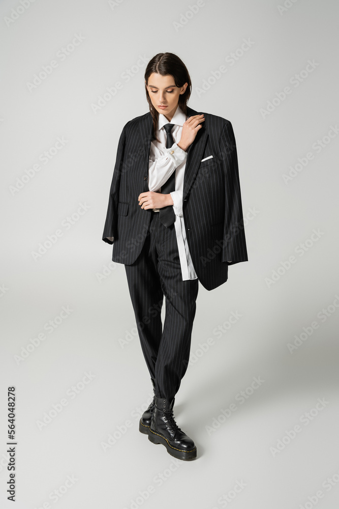 full length of young woman in black elegant suit and rough leather boots looking down on grey background. full length of young woman in black elegant suit and rough leather boots looking down on grey background.