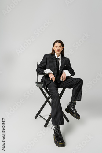full length of trendy woman in rough boots and black oversize suit sitting on chair and looking at camera on grey background.