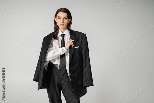 brunette model in white shirt and black trousers with blazer looking at camera isolated on grey.