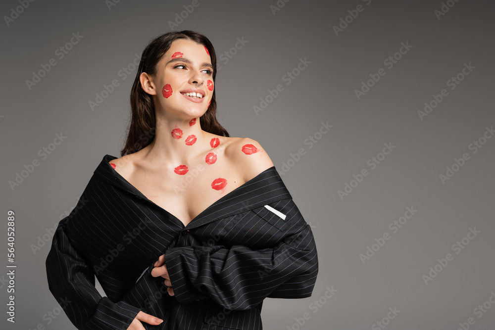 cheerful young woman with red kisses on face and body looking away while posing in oversize blazer isolated on grey. cheerful young woman with red kisses on face and body looking away while posing in oversize blazer isolated on grey.