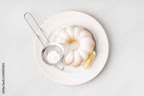 Plate with delicious cake, sieve of powdered sugar and lemon slice on white background