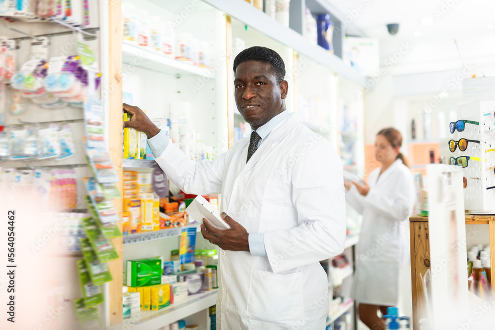 Concentrated african american male pharmacist working in a pharmacy is standing near a rack, laying out goods on the shelves