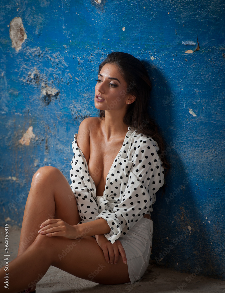 Beautiful girl with white shirt and white shorts posing in old hall against blue wall. Attractive long hair brunette side view against ancient pillars. Dark hair young woman with gorgeous eyes