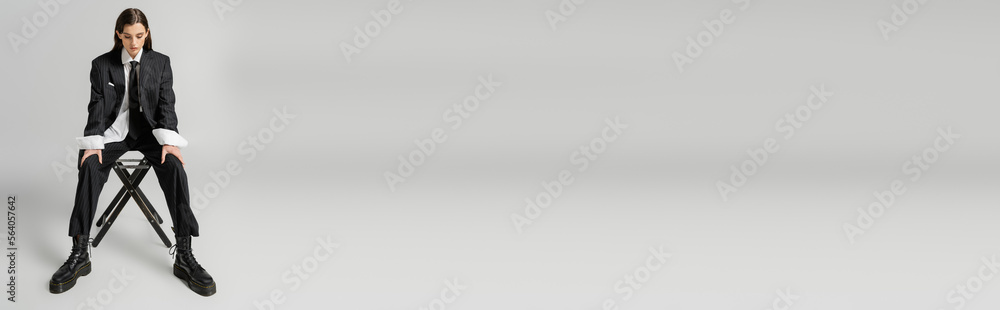 Φωτογραφία full length of stylish woman in elegant pantsuit and rough boots sitting on chair on grey background, banner