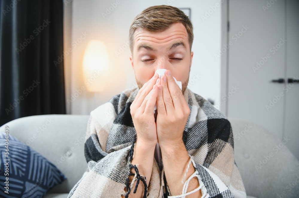 Person with cold blowing his nose. Sick man wearing scarf sitting on couch at home. Man with red running nose and cough Person with cold blowing his nose. Sick man wearing scarf sitting on couch at home. Man with red running nose and cough