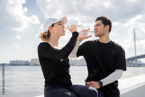 A sporty man and a woman drink mineral water with vitamins from a bottle, a break after running.