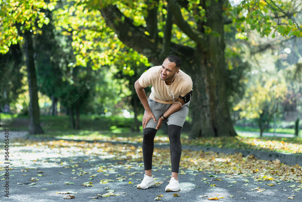 Runner injured his leg while jogging in the park near the trees, hispanic man has muscle spasm massaging his knee, man is running and exercising fitness on a sunny day. Runner injured his leg while jogging in the park near the trees, hispanic man has muscle spasm massaging his knee, man is running and exercising fitness on a sunny day.