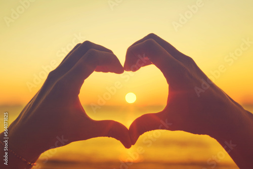 Women's hands in the shape of a heart against the background of the sea and the setting sun let the sun's rays through, toned. Hands in the shape of a heart of love. Romantic background