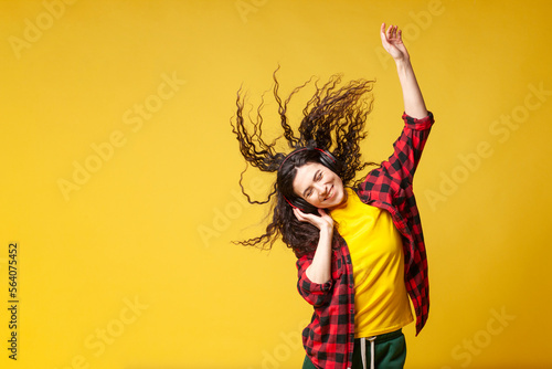 young curly brunette girl in a plaid shirt listens to music in headphones and dances with raised hands