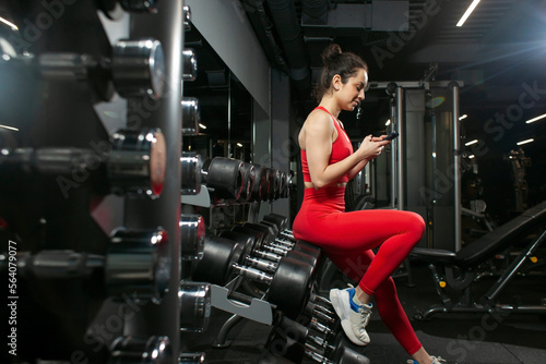 young sporty girl in red sportswear is using smartphone in the gym and typing a message, attractive slim woman