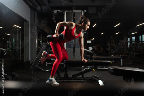 young sports girl in red sportswear lifts heavy dumbbells and does exercise in the gym, athletic woman trains