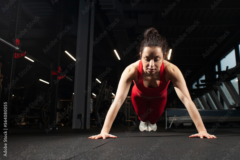 Fototapeta premium young athletic girl in red sportswear is doing push-ups from the floor in a black gym, an attractive woman is training