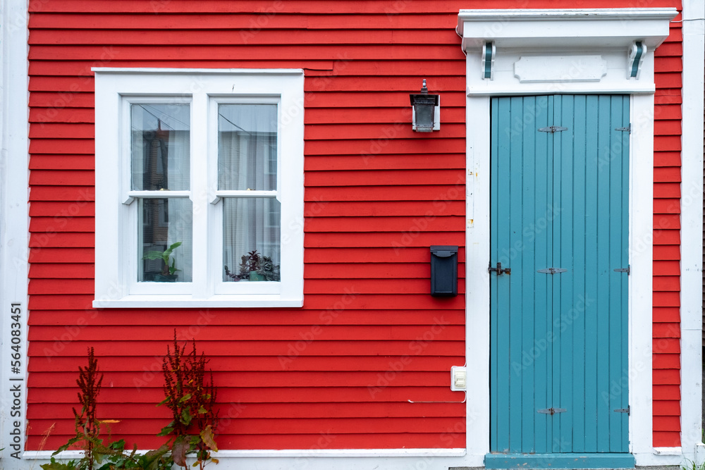 Fototapeta premium A bright red wooden exterior wall of a country style house. The building has red narrow wood clapboard cape cod siding. It has a small wooden door, a double pane glass window, and a black mailbox.