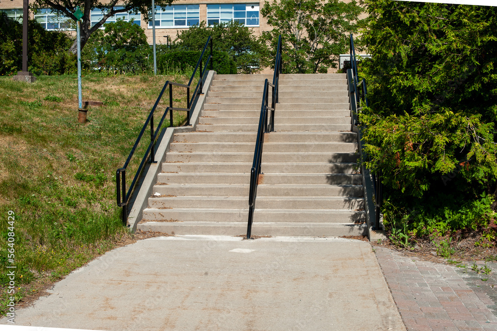 Concrete steps lead to a basketball court. There are black metal handrails dividing the two stairways to the outside park area. The grounds have trees, grass and brick building on the top level.