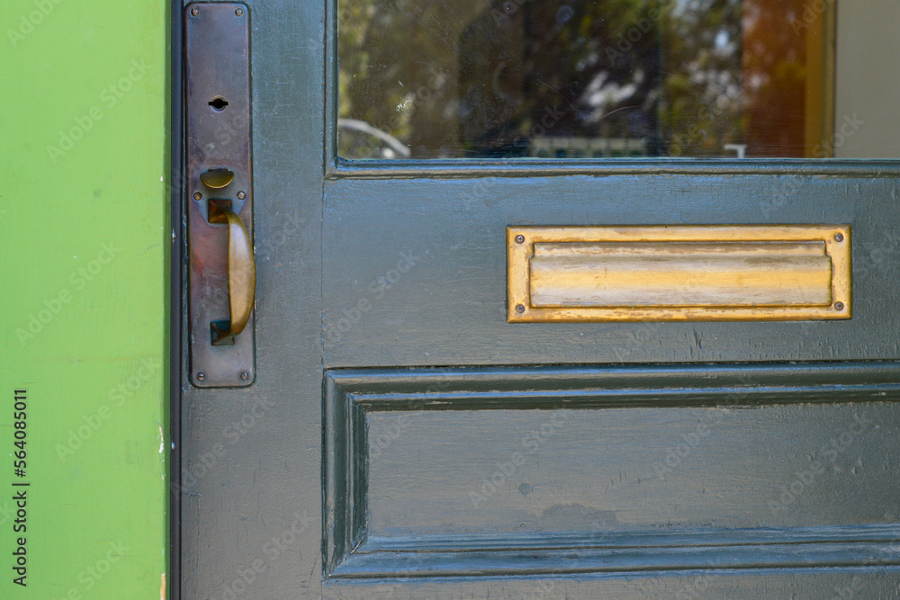 A dark green wooden door with half glass pane window, and a wooden panel. The vintage door has a brass door handle, a keyhole, and a letter plate. The trim of the building is vibrant green color. 