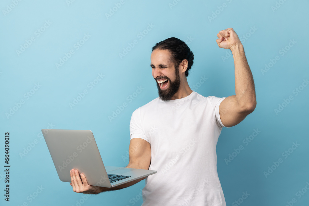 Portrait of successful man with beard wearing white T-shirt screaming with joy and holding laptop, rejoicing victory, online betting. Indoor studio shot isolated on blue background. Portrait of successful man with beard wearing white T-shirt screaming with joy and holding laptop, rejoicing victory, online betting. Indoor studio shot isolated on blue background.