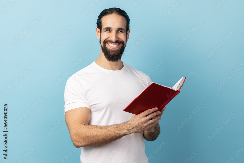 Portrait of man with beard wearing white T-shirt holding book with happy facial expression, reading, standing looking at camera with toothy smile. Indoor studio shot isolated on blue background. Portrait of man with beard wearing white T-shirt holding book with happy facial expression, reading, standing looking at camera with toothy smile. Indoor studio shot isolated on blue background.