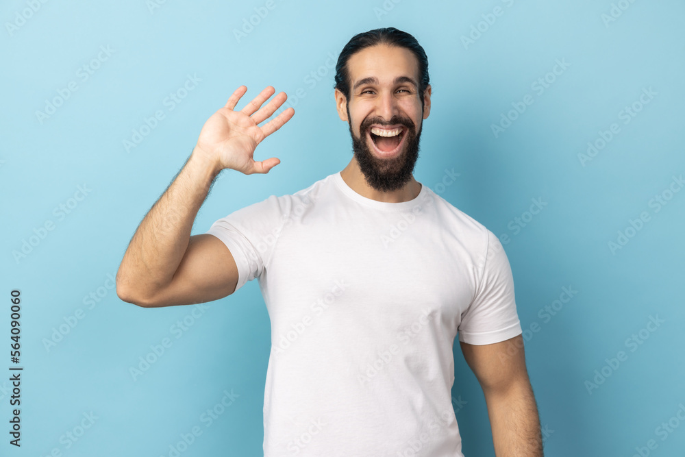 Portrait of positive man with beard wearing white T-shirt standing with raised palm gesturing hi to camera, welcoming with toothy smile. Indoor studio shot isolated on blue background. Portrait of positive man with beard wearing white T-shirt standing with raised palm gesturing hi to camera, welcoming with toothy smile. Indoor studio shot isolated on blue background.