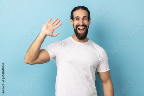 Portrait of positive man with beard wearing white T-shirt standing with raised palm gesturing hi to camera, welcoming with toothy smile. Indoor studio shot isolated on blue background.