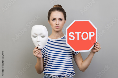 Portrait of serious woman wearing striped T-shirt holding white mask with unknown face and red traffic sign, looking at camera. Indoor studio shot isolated on gray background. photo