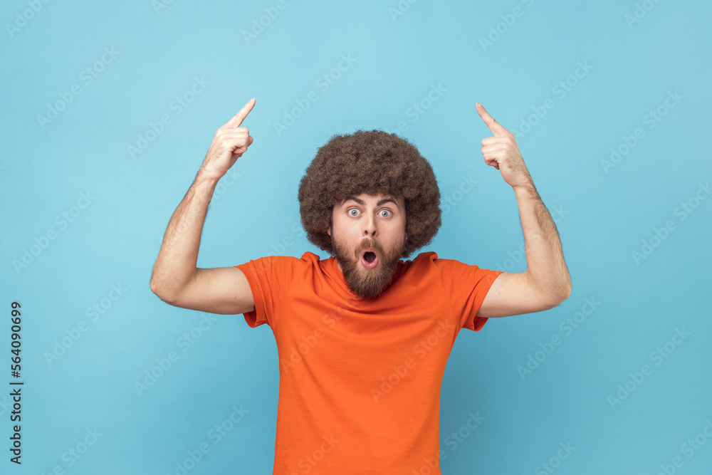 © khosrork - Wow, look, crazy advertisement. Man with Afro hairstyle in orange T-shirt pointing up at empty place for ad content and expressing astonishment. Indoor studio shot isolated on blue background.