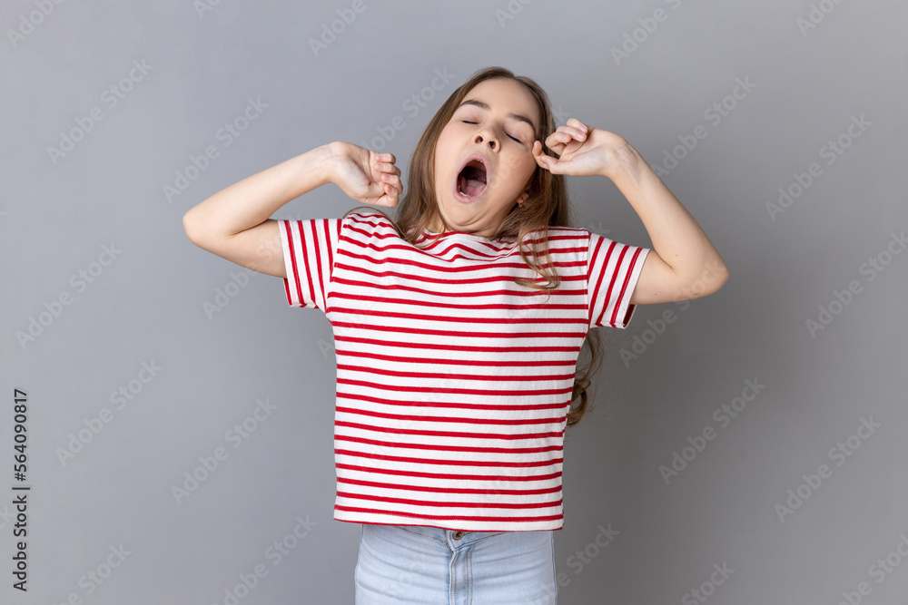 Portrait of sleepy little girl wearing striped T-shirt yawning and raising hands up, feeling fatigued, standing with close eyes. Indoor studio shot isolated on gray background. Portrait of sleepy little girl wearing striped T-shirt yawning and raising hands up, feeling fatigued, standing with close eyes. Indoor studio shot isolated on gray background.