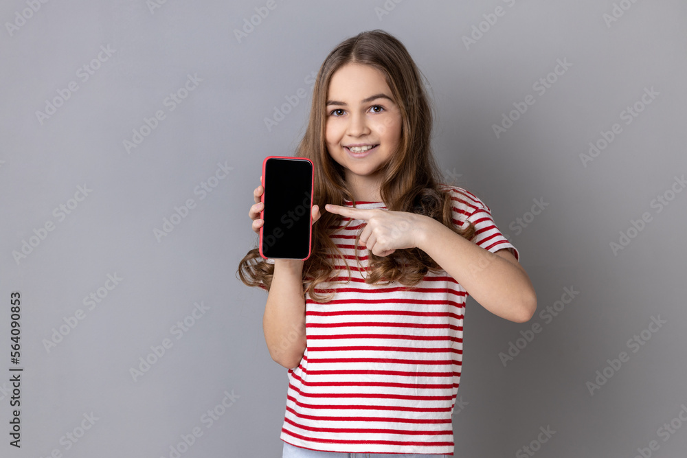 Portrait of delighted little girl wearing striped T-shirt pointing finger at smartphone with white empty display, bragging with application. Indoor studio shot isolated on gray background. Portrait of delighted little girl wearing striped T-shirt pointing finger at smartphone with white empty display, bragging with application. Indoor studio shot isolated on gray background.