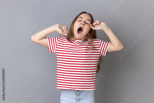 Portrait of sleepy little girl wearing striped T-shirt yawning and raising hands up, feeling fatigued, standing with close eyes. Indoor studio shot isolated on gray background. photo