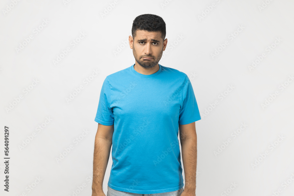 Portrait of sad upset depressed unshaven man wearing blue T- shirt standing looking at camera with frowning face, expressing sadness, being offended. Indoor studio shot isolated on gray background. Portrait of sad upset depressed unshaven man wearing blue T- shirt standing looking at camera with frowning face, expressing sadness, being offended. Indoor studio shot isolated on gray background.